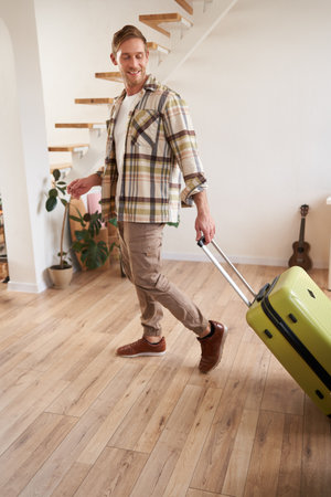 Vertical portrait of happy young man with suitcase, walking indoors, leaving a flat with luggage, going on business trip or vacationの写真素材