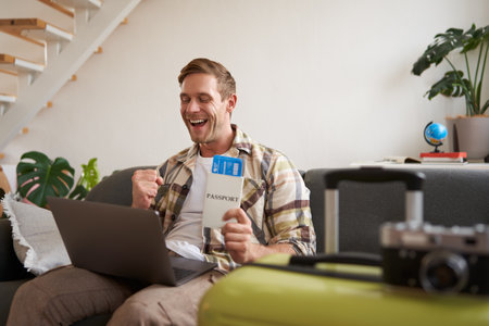 Portrait of young handsome man sitting with laptop, has passport and two plane tickets prepared, planning his journey on computer, booking hotels and restaurants online for the tripの写真素材