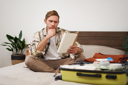 Portrait of man with suitcase, sits in room and looking serious at his notes, packing for holiday, checking list of items in notebookの写真素材