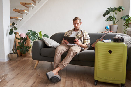 Image of young man with tablet, sitting in living room with suitcase, reading, studying the route, preparing for a vacationの写真素材
