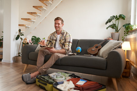 Portrait of smiling man with suitcase, looks at his notes in notebook, checks list of items for holidayの写真素材
