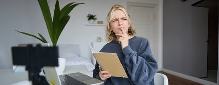 Portrait of young woman, student sitting in room, studying from home, remote education concept, holding notebook and thinking, looking aside with thoughtful face, concentrating on assignmentの写真素材