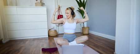 Portrait of young athletic woman doing workout at home, shows her muscles, strong biceps, sits on yoga mat and wears wireless headphonesの写真素材