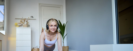 Vertical shot of young blond woman at home, wearing wireless headphones, looking at laptop, standing in plank, watching fitness tutorial onlineの写真素材