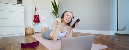 Young woman doing fitness workout at home, lying on rubber yoga mat, holding smartphone, wearing headphones, using laptop to watch sports videosの写真素材