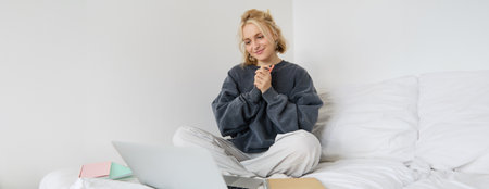 Portrait of young beautiful woman sitting on bed with laptop and notebooks, working from home, freelancing. Female student studying online, video chats, connects to lesson remotelyの写真素材