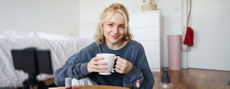Close up portrait of beautiful blond woman, girl sits in her room, does makeup, records video for social media, drinks tea, holds mug and smiles at cameraの写真素材