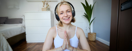 Portrait of young relaxed woman, sits in room in headphones, clasp hands together, meditated, listens to yoga musicの写真素材