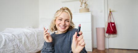 Portrait of young social media influencer, woman recording a video with beauty products, showing her makeup on camera, holding lipstick and smiling, sitting on floorの写真素材