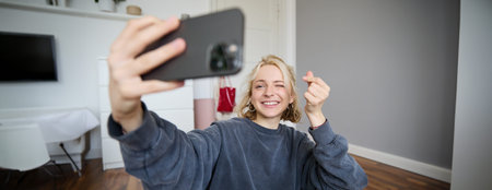 Portrait of young stylish girl sits on bedroom floor, takes selfies on her smartphone, posing for...
