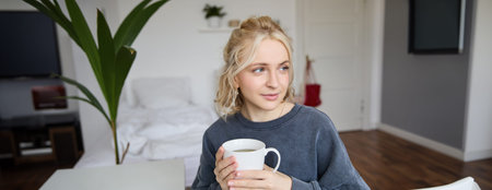 Close up portrait of smiling young blond woman, sits with cup of tea in bedroom, rests at home,...