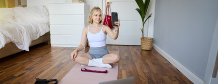 Portrait of stylish young woman, sitting on rubber mat on floor at home, taking selfie while doing workout, yoga session, posting photo on social mediaの写真素材