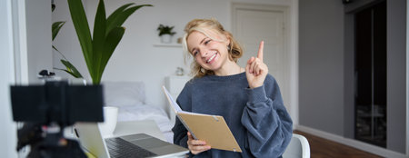 Portrait of young smiling, charismatic woman, raising one finger, teaching online, reading from notebook, looking at laptop, recording video course, content maker working on new project from homeの写真素材