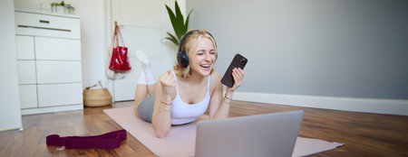 Portrait of young woman workout, watching exercise videos on laptop in headphones, lying on rubber mat with mobile phone and smilingの写真素材