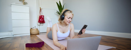 Portrait of happy, smiling sporty woman, doing workout at home, using laptop to watch workout videos in headphones, checking sports app on mobile phoneの写真素材