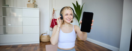 Portrait of fit and healthy woman workout at home, wearing headphones, showing smartphone screen, empty dark displayの写真素材