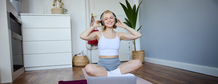 Portrait of young fitness girl, woman workout at home, listens to fitness instructor in wireless headphones, follows exercises on laptopの写真素材