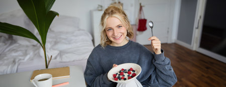 Portrait of smiling blond woman, eating breakfast, holding bowl and spoon, sitting in bedroom, looking happy at cameraの写真素材