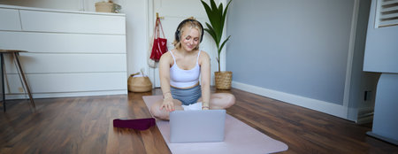Portrait of woman during workout, sitting on yoga mat with resistance band, listening to video instructions on laptop, wearing wireless headphones, repeating exercisesの写真素材