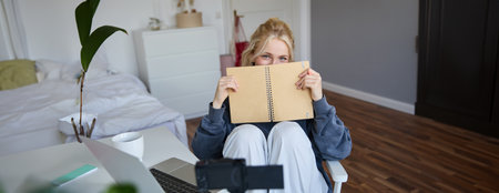 Portrait of cute blond woman, sits in front of digital camera and laptop in her room, covers face with notebook journal, records video of herself for lifestyle vlogの写真素材