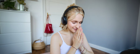 Portrait of young relaxed woman, sits in room in headphones, clasp hands together, meditated, listens to yoga musicの写真素材