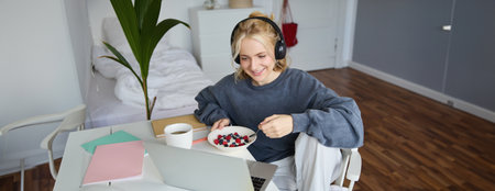 Portrait of happy young blond woman, sitting in a room, watching movie on laptop and eating healthy breakfast, drinking tea, resting on weekendの写真素材