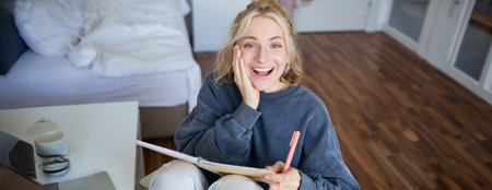 Portrait of happy, young blond woman, sitting in bedroom with notebook and pen, laughing and smiling, writing in journal, doing homework and smilingの写真素材