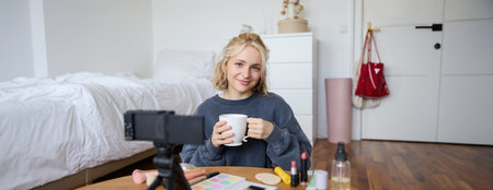 Portrait of beautiful social media beauty blogger, sitting in front of digital camera on floor in bedroom, drinking tea and chatting, talking to followersの写真素材