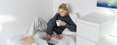Portrait of young woman, student studying in her bed, relaxing while preparing homework, eating doughnut, using laptop in bedroom and drinking teaの写真素材