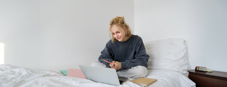 Portrait of young smiling woman studying in her bed, working from home in bedroom, sitting with laptop and notebooks on lotus pose, looking happy and relaxedの写真素材