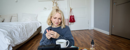Close up portrait of young lifestyle blogger, woman sits in front of camera, looks thoughtful, applies makeup, holds mascara, explains how to put on beauty product for social media followersの写真素材