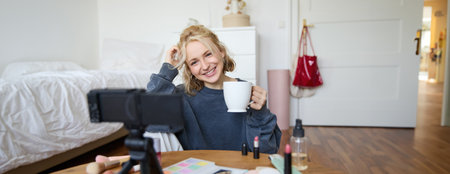 Portrait of blond young lifestyle blogger, woman records video of her talking about life and beauty, sits in front of camera, holds cup, drinks tea, does blog content for social media accountの写真素材