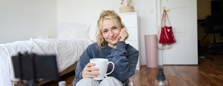 Close up portrait of beautiful blond woman, girl sits in her room, does makeup, records video for social media, drinks tea, holds mug and smiles at cameraの写真素材