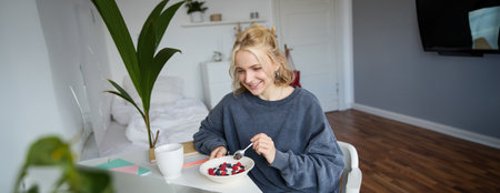 Portrait of smiling blond young woman, eating in front of laptop, watching videos online while having breakfast, enjoying dessert, sitting in bedroomの写真素材