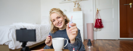 Portrait of smiling woman shows lip gloss, advertising makeup on her social media account, recording video on digital camera, content maker working on new project in her roomの写真素材