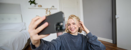 Portrait of young woman, social media influencer, taking selfies in her room, sitting on floor, holding smartphone and posing for a photoの写真素材