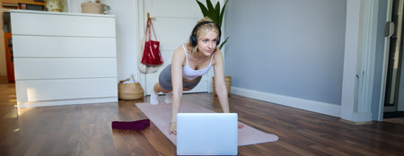 Portrait of young athletic woman in wireless headphones, standing in plank, following online workout video, using laptop to join training session on the internetの写真素材