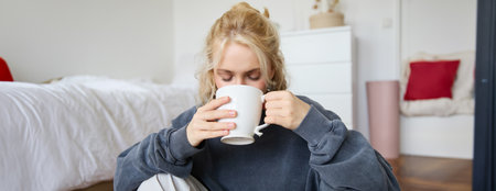 Portrait of young woman sitting on bedroom floor, drinking tea, holding white mug and smiling at cameraの写真素材