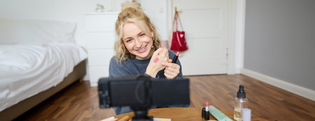 Image of young stylish woman, blogger recording a beauty lifestyle video of her picking best lipstick, showing lip balm swatches on her skin, sitting in front of digital camera in empty roomの写真素材