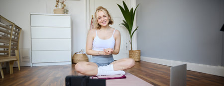 Young female athlete, fitness instructor woman sits on floor rubber mat, recording video on digital camera, showing how to workout, explaining exercisesの写真素材