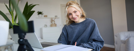 Portrait of beautiful smiling woman, student studying at home, remote education concept, sits in a room with laptop and writes down notes, uses her journalの写真素材