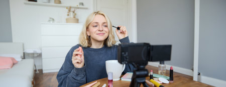 Image of cheerful, beautiful young lifestyle blogger, woman sitting on floor and recording video about makeup, holding mascara, making lifestyle content for her social media account and followersの写真素材