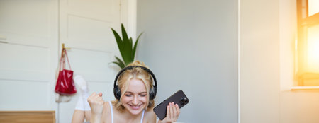 Vertical shot of young fitness woman in headphones, lying on rubber mat, relaxing after workout training session, holding smartphone, smiling at cameraの写真素材