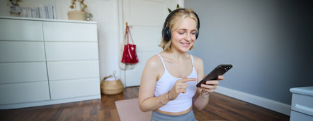 Portrait of young sporty woman in wireless headphones, sitting with smartphone on rubber mat, using workout training app, fitness application on her phone, listening in earphonesの写真素材