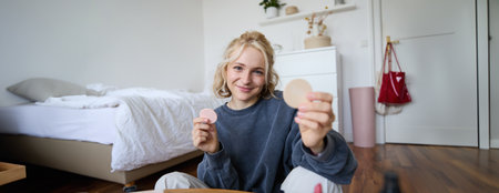 Portrait of young woman, content creator, showing beauty makeup products at camera, sitting on floor in bedroom and smilingの写真素材