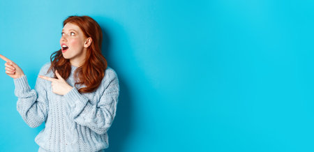 Winter holidays and people concept. Amazed redhead girl checking out promo offer, pointing and looking left at copy space, standing against blue backgroundの写真素材