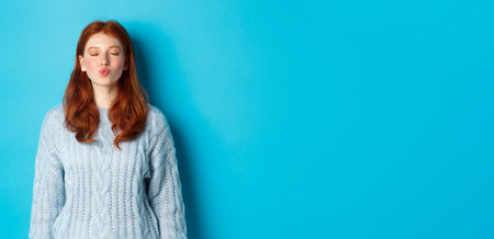 Cute redhead teen girl waiting for kiss, pucker lips and close eyes, standing in sweater against blue backgroundの写真素材