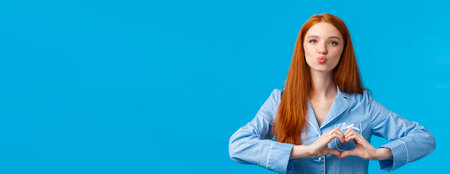 Lovely sensual good-looking redhead female with long ginger hair, freckles, folding lips for kiss showing heart love gesture over chest, wearing nightwear, ready sleep, blue backgroundの写真素材