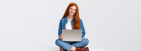Cheerful sassy redhead female student, college girl in glasses, sitting on crossed legs with laptop, working freelance, prepare project awat from office, remote over white backgroundの写真素材