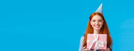 Waist-up portrait cheerful happy, pretty redhead, ginger girl celebrating birthday, receive cute pink wrapped present, wearing b-day cap and smiling, having fun at party, blue backgroundの写真素材